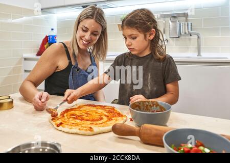 Mutter schaut auf Tochter Anwendung Sauce auf Pizzateig in Küche Stockfoto