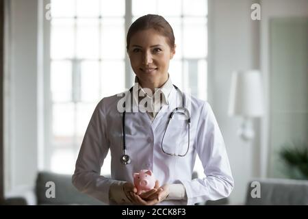 Lächelnd angenehme junge Ärztin hält Sparschwein in den Händen. Stockfoto