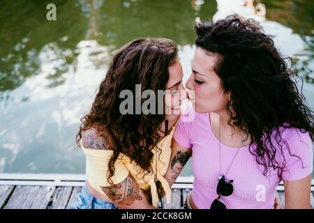 Nahaufnahme einer jungen Frau, die sich während des Sitzens auf die Nase der Freundin küsst Am See Stockfoto