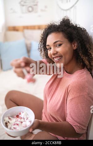 Fröhliche Frau, die Erdbeeren mit Sahne zeigt, während sie auf dem Bett sitzt Zu Hause Stockfoto