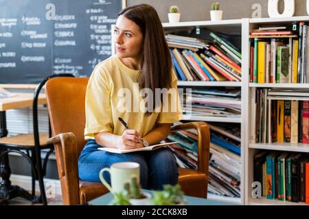 Nachdenkliche Frau, die im Buch schreibt, während sie auf einem Stuhl sitzt Café-Bar Stockfoto