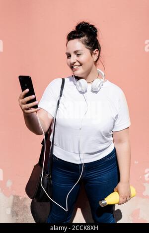 Lächelnd kurvige junge Frau mit Handy und Kopfhörer gelehnt Steht an einer Wand Stockfoto