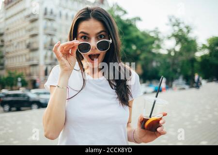 Überrascht junge Frau mit Mund offen trägt Sonnenbrille im Stehen An der City Street Stockfoto