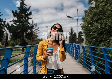 Fröhliche Frau trägt eine Sonnenbrille und spricht im Stehen über ein Smartphone Auf dem Steg im Park Stockfoto