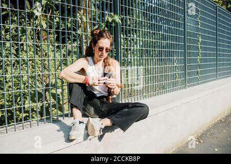 Frau, die Mobiltelefon benutzt, während sie auf einer Stützwand sitzt Zaun Stockfoto