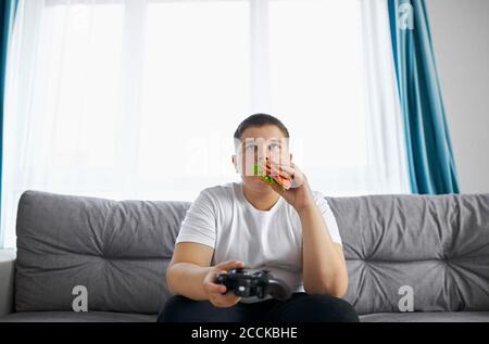 Keen kaukasischen Jungen spielen Videospiele, übergewichtigen Jungen sitzen auf dem Sofa allein, hat Probleme mit der Ernährung, führen ungesunden Lebensstil. Faulheit Konzept Stockfoto