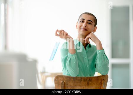 Porträt einer selbstbewussten Frau auf einem Stuhl in einem Loft hält schützende Gesichtsmaske Stockfoto