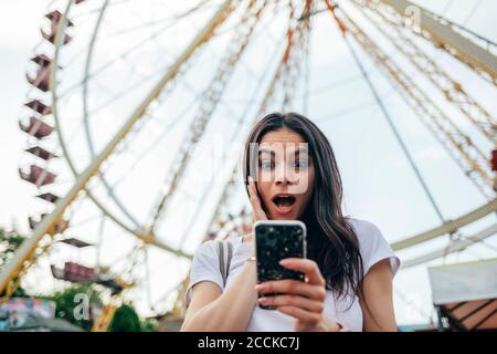 Überrascht junge Frau mit Mund offen mit Smartphone gegen Riesenrad im Vergnügungspark Stockfoto