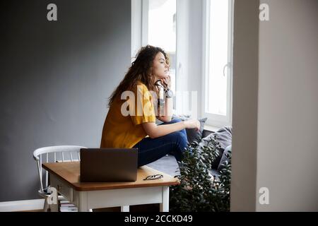 Nachdenkliche Frau, die durch das Fenster schaut, während sie Pause von der Arbeit nimmt Im Wohnzimmer im Home Office Stockfoto