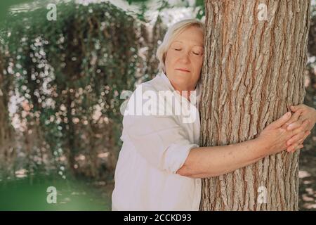 Ältere Frau mit geschlossenen Augen umarmt Baumstamm im Stehen Im Hof Stockfoto