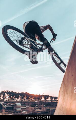 Sorgloser junger Mann, der Stunt mit Fahrrad auf Rampe gegen Himmel im Park bei Sonnenuntergang Stockfoto