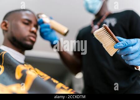 Barbier trägt Handschuhe, die die Bürste halten, während er die Haare des jungen Mannes schneidet Im Salon Stockfoto