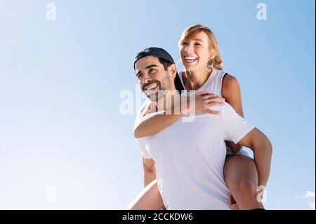 Mitte Erwachsene Mann Huckepack fröhliche Frau gegen klaren Himmel während Sonniger Tag Stockfoto