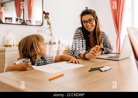 Lächelnde Mutter mit Laptop, die Tochter beim Malen auf dem Tisch ansieht Im Esstisch Stockfoto