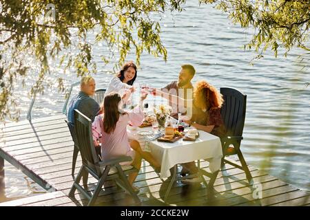 Freunde, die auf dem Steg an einem See zu Abend essen und Wein anklirren Brille Stockfoto