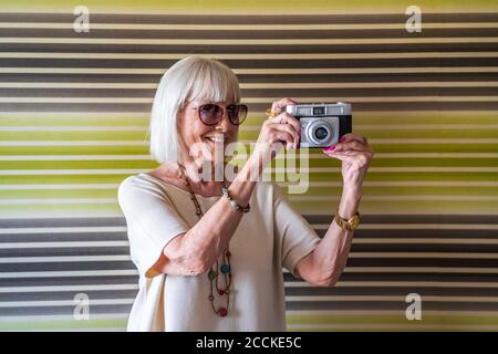 Lächelnde ältere Frau mit Sonnenbrillen, die mit der Kamera gegen die Wand fotografieren Zu Hause Stockfoto