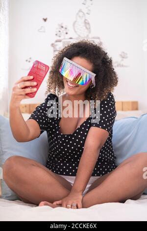 Frau trägt mehrfarbige Sonnenbrille, die Selfie mit Smartphone nimmt Auf dem Bett zu Hause Stockfoto