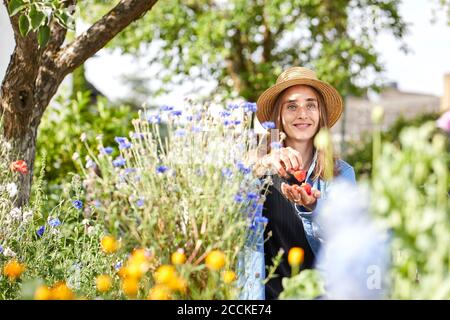 Lächelnde junge Frau, die Erdbeere isst, während sie inmitten von Pflanzen sitzt Garten Stockfoto
