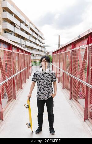 Junger Mann hält Skateboard, während er auf einem Steg in der Stadt steht Stockfoto