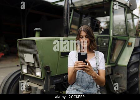Junge Frau mit Handy an einem Traktor auf einem Bauernhof Stockfoto