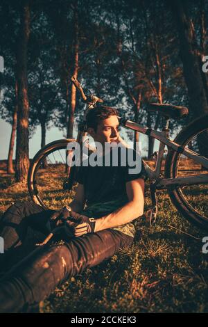 Nachdenklicher junger Mann, der während des Sonnenuntergangs mit dem Fahrrad im Park sitzt Stockfoto