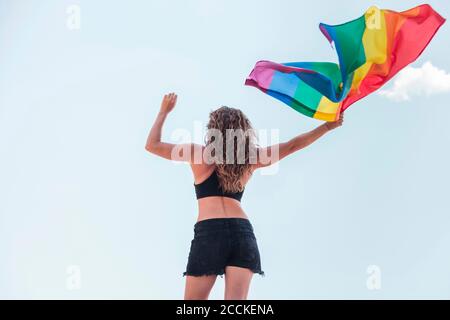 Frau mit mittlerem Erwachsenen, die Regenbogenfahne schwenkt, während sie gegen den Himmel steht Stockfoto