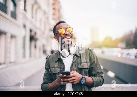 Fröhlicher Mann mit Sonnenbrille und Smartphone im Stehen Stadt Stockfoto