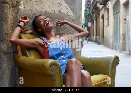 Fröhliche Frau mit erhobenen Armen sitzt auf alten Sessel in Straße Stockfoto
