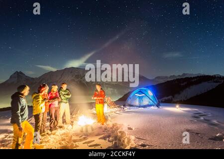 Nachtsicht von glühendem Zelt im Schnee und Menschen um Feuer, Achenkirch, Österreich Stockfoto