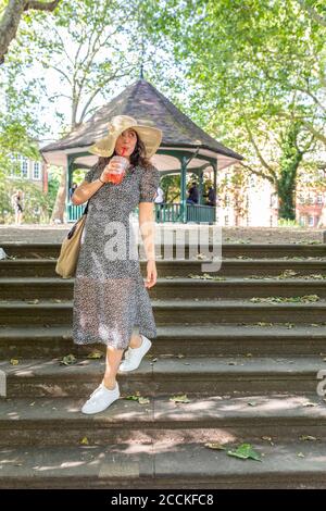 Junge Frau mit Hut trinken, während sie sich auf Treppen im Park Stockfoto