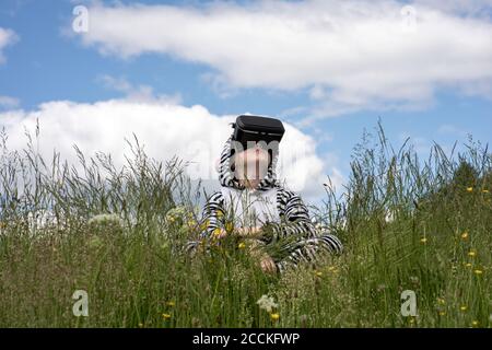 Junge trägt Zebra-Kostüm mit VR-Simulator, während sitzend auf Gras am Himmel Stockfoto