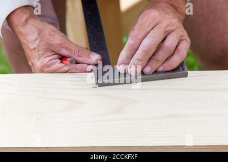 Nahaufnahme der Hände eines älteren Handwerkers - Zimmermann bei der Arbeit mit einem Metalllineal und einem grafischen Bleistift Zeichnung Markierungen auf einem Holzbalken. Stockfoto