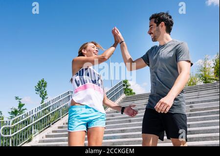 Fröhliches Paar, das High-Five gibt, während es auf Stufen gegen Blau steht Himmel in der Stadt Stockfoto