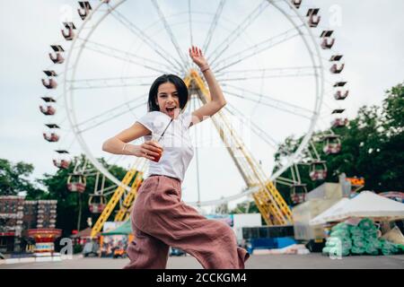 Junge Frau hält Softdrink Tasse tanzen gegen Riesenrad Im Vergnügungspark Stockfoto