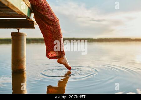 Frau, die auf dem Steg an einem See bei Sonnenuntergang sitzt Das Wasser mit dem Fuß Stockfoto