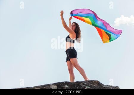 Frau mittleren Erwachsenen winkt Regenbogenfahne, während sie auf dem Felsen steht Gegen den Himmel Stockfoto