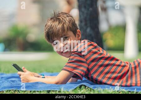 Lächelnder Junge, der Smartphone benutzt, während er auf einem Handtuch liegt Hof Stockfoto