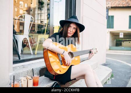 Schöne Frau trägt Hut Gitarre spielen, während draußen Café sitzen In der Stadt Stockfoto