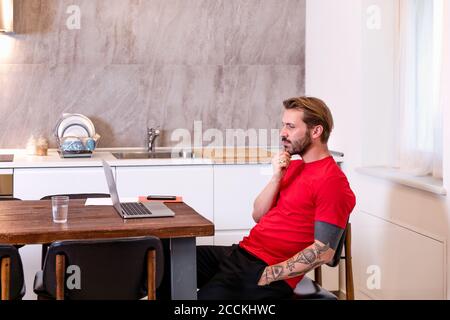 Nachdenklicher Mann mittleren Erwachsenen, der auf den Laptop auf dem Esstisch schaute Während Sie in der Küche sitzen Stockfoto