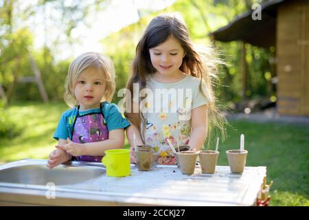 Nettes Mädchen stehen mit Schwester Pflanzen Samen in kleinen Töpfen Auf dem Tisch im Hof Stockfoto