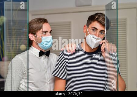 Der junge Mann mit einer OP-Maske legt seine Hand auf die Schulter eines anderen jungen Mannes, der mit einer Gesichtsmaske telefoniert. Mitarbeiterkonzept. Stockfoto