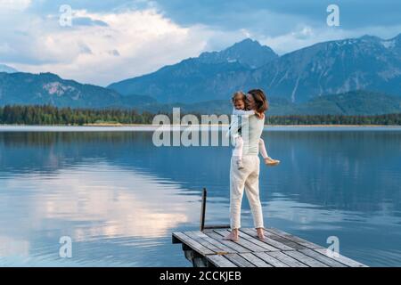 Glückliche Mutter, die Tochter trägt, während sie auf dem Steg über dem See steht Gegen den Berg Stockfoto