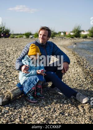 Lächelnder Vater mit Tochter, die am Ufer auf dem Balken sitzt Himmel an sonnigen Tagen Stockfoto