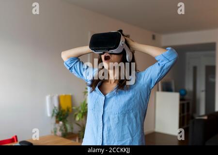 Junge Frau mit Mund offen Blick durch virtuelle Realität Simulator Im Heimbüro stehen Stockfoto