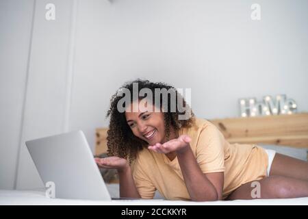 Fröhliche junge Frau Videokonferenzen über Laptop, während sie auf liegt Bett zu Hause Stockfoto