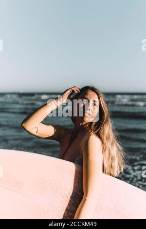 Junge Frau, die Surfbrett trägt, schaut weg, während sie gegen das Meer steht Stockfoto