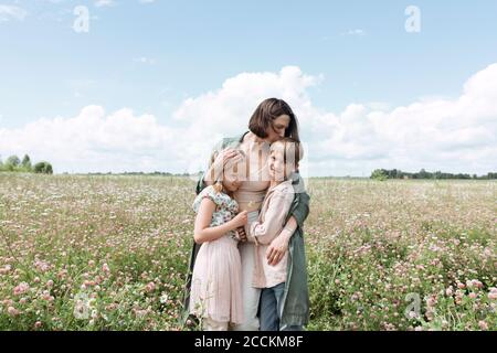 Liebevolle Mutter umarmt Kinder, während sie inmitten von Blumen auf dem Feld steht Gegen den Himmel Stockfoto