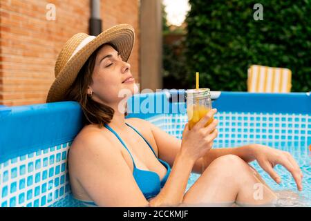 Frau hält Orangensaft, während sie sich im aufblasbaren Swimmingpool entspannt Am Hof Stockfoto