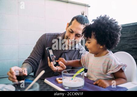 Lächelnder Vater, der der kleinen Tochter beim Essen ein Smartphone zeigt Tabelle Stockfoto