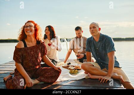 Glückliche Freunde, die am Steg an einem See picknicken Sonnenuntergang Stockfoto